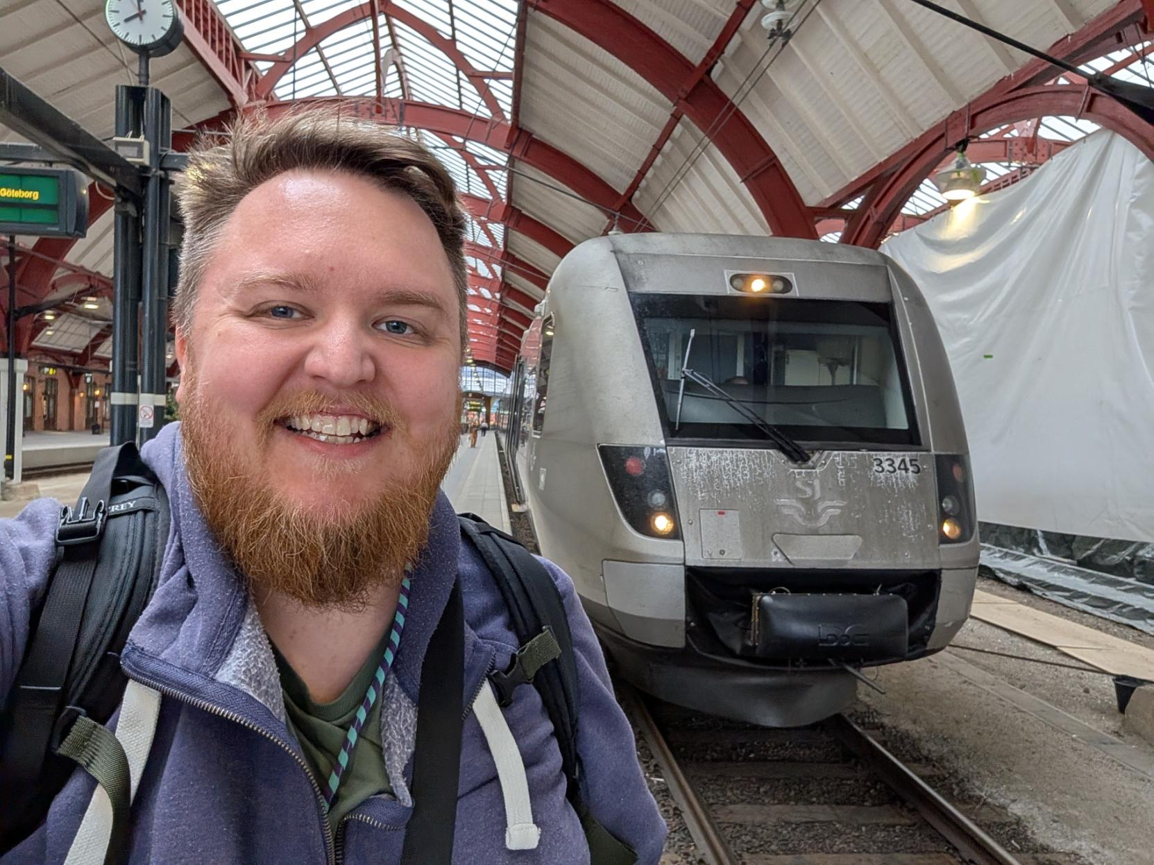 Ein selfie av ein breitt smilande mann med skjegg og lilla hettegenser på ein togperrong inne på Malmö Centralstation. Til høgre står den grå, strømlinjeforma fronten på eit svensk SJ X2000-snøggtog på sporet. Togfronten er prega av metalloverflata, har SJ sin klassiske, vinga logo prega i midten, og vognnummeret «3345» oppe til høgre. Over og bak toget breier det historiske, raudmåla perrongtaket i stål og tre seg ut. Oppe til venstre ser ein ei analog stasjonsklokke og eit digitalt skilt der det står «Göteborg».