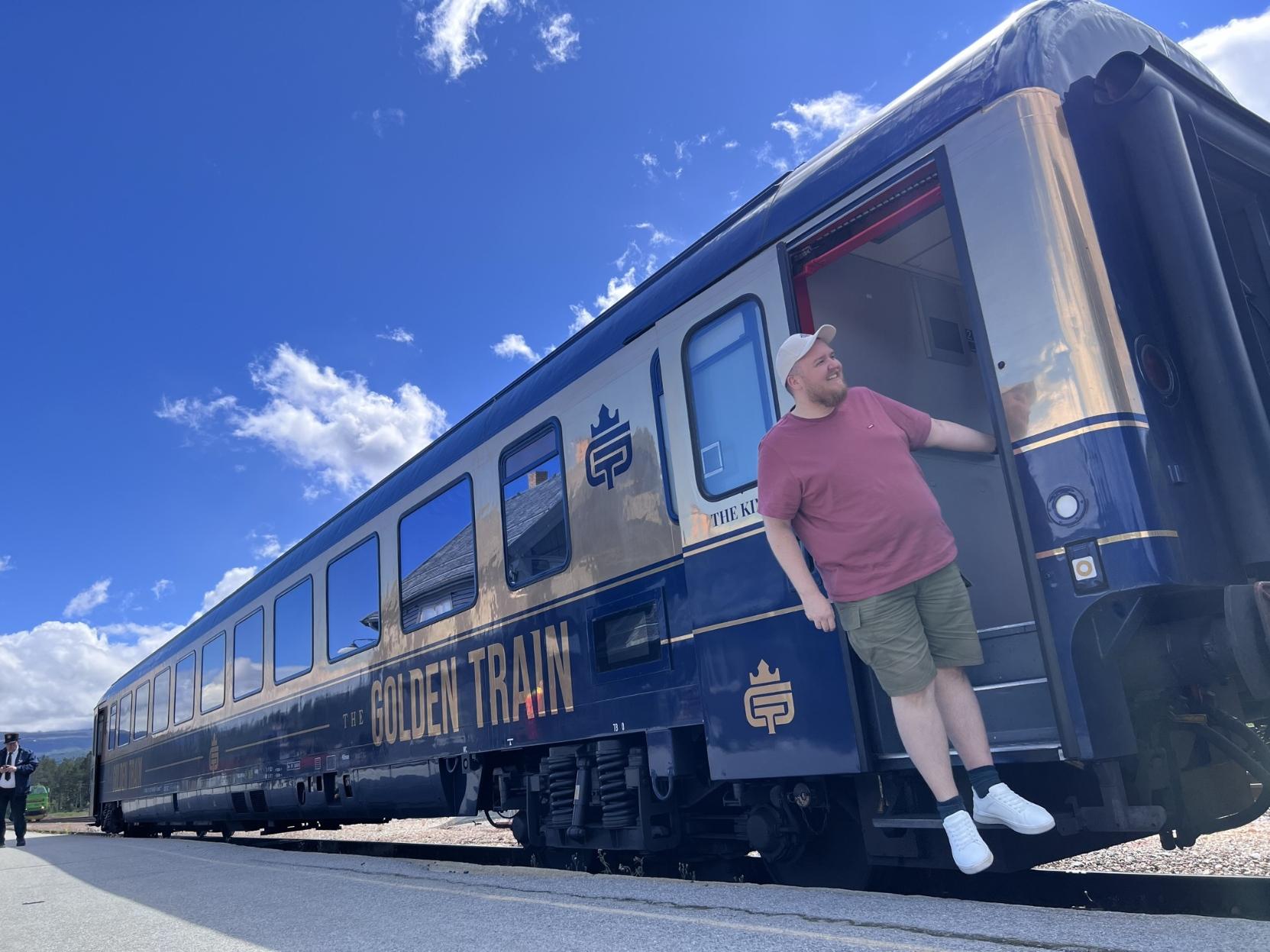 Eit bilete teke i lågt perspektiv frå ein stasjonperrong i klårt solskin, som viser den bakre delen av eit passasjertog frå The Golden Train. Ein smilande mann med skjegg, ljos caps, rosa t-skjorte og shorts heng spøkefullt ut frå den opne døra heilt bakarst på vogna medan han held seg fast i dørkarmen. Togvogna er elegant lakkert i mørkeblått med eit breitt gullfarga felt langs vindaugsrekkja, og har teksten «THE GOLDEN TRAIN» og ein kongekrone-logo i gull på vognsida. Toget strekkjer seg framover til venstre i biletet under ein klår, sterk blå himmel med nokre få, kvite skyer. Ein uniformert togkonduktør kan skimtast på perrongen langt til venstre.