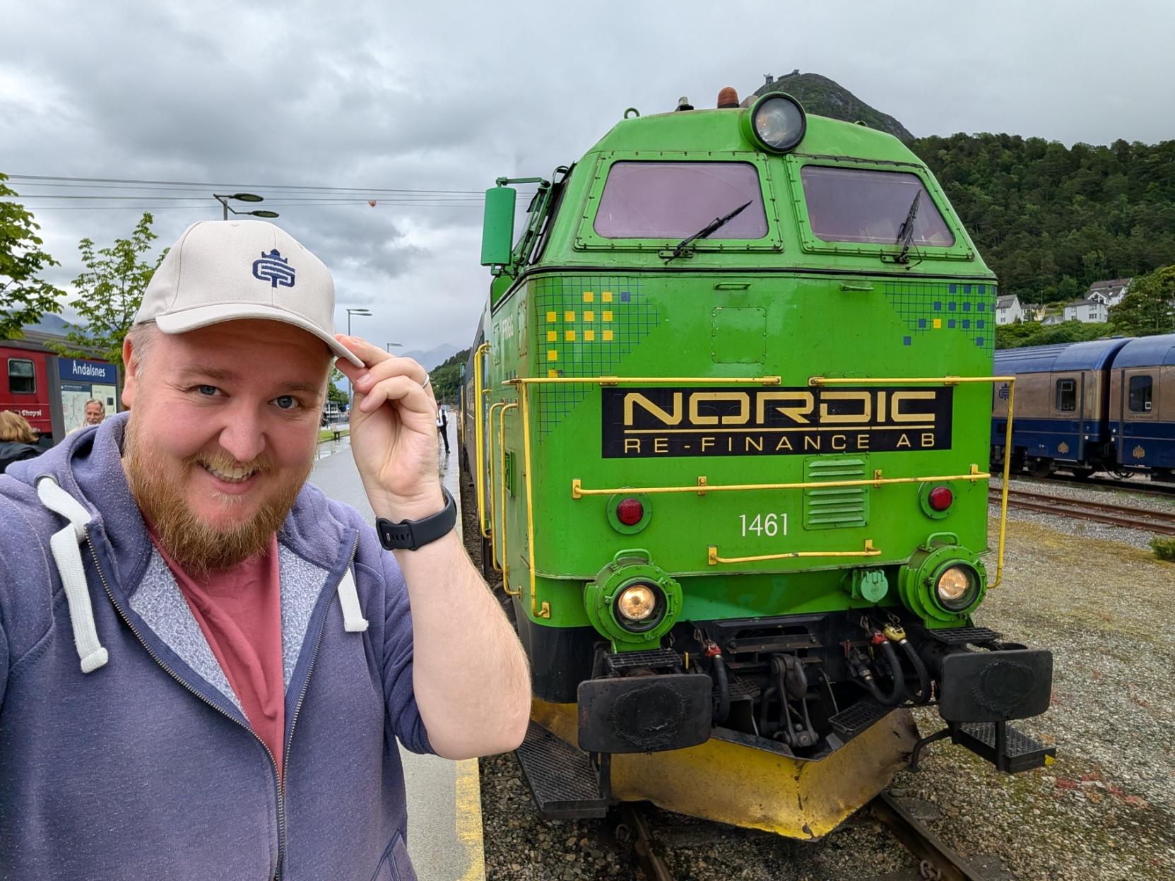 Ein selfie av ein smilande mann med skjegg, lilla hettegenser og ein lys caps som han helsar med, på ein perrong ved Åndalsnes stasjon. Til høgre dominerer den flate, kantete fronten på eit kraftig, lysegrønt diesellokomotiv som dreg The Golden Train. Midt på den grøne fronten er det eit stort, svart skilt med gullskrift der det står «NORDIC RE-FINANCE AB», og under dette står lokomotivnummeret «1461». Lokomotivet har frontlysa tende. I bakgrunnen til høgre kan ein sjå dei mørkeblå og gullfarga passasjervognene til The Golden Train oppstilt på sporet ved sidan av, og bakom reiser det grønkledde fjellet Nesaksla seg under ein overskya himmel.