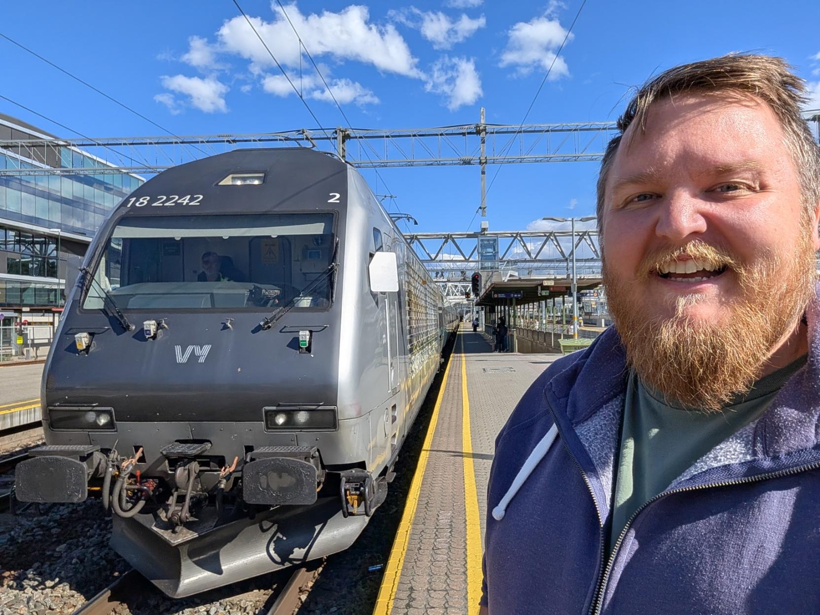 Ein selfie av ein breitt smilande mann med skjegg, grøn t-skjorte og blå hettejakke på ein solfylt perrong på Oslo Sentralstasjon (Oslo S). Til venstre står fronten på eit mørkegrått elektrisk lokomotiv av typen El 18, som dreg toget over Bergensbanen mot Bergen. Lokomotivfronten har det karakteristiske, hellande designet og er merka med lokomotivnummeret «18 2242» øvst over frontruta, med eit lite «2» oppe til høgre. Midt på snuten prydar den kvite Vy-logoen, og i førarrommet skimtar ein lokføraren bak frontruta. Bak lokomotivet strekkjer vognrekkja seg innover langs perrongen. I bakgrunnen ser ein fagverksåk med køyreleidningar over spora, glasbygg til venstre og eit signal i raudt, alt under ein klår, blå himmel med nokre få, kvite skyer.

🚆 Tog: El 18 · Nr: 18 2242 · Stasjon: Oslo S · Bane: Bergensbanen · Selskap: Vy