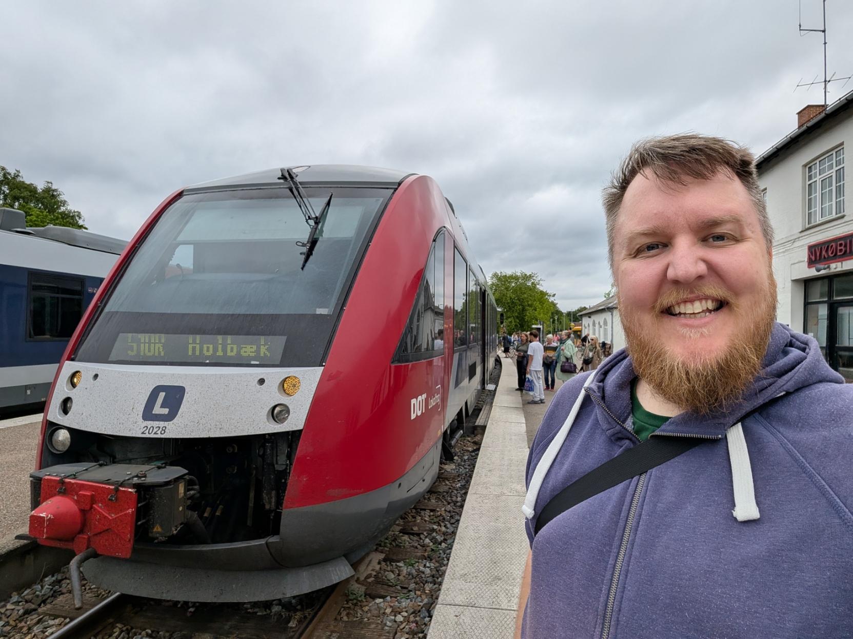 Ein selfie av ein breitt smilande mann med skjegg og lilla hettegenser på Nykøbing Sjælland stasjon. Til venstre står fronten på eit raudt og grått lokaltog av modellen Siemens Desiro Classic på sporet. Det digitale destinasjonsskiltet i frontruta viser teksten «510R Holbæk». Til høgre i biletet ser ein stasjonsbygningen i mur med eit skilt der det står «NYKØBING». I bakgrunnen på den grå perrongen står det fleire andre reisande som ventar under ein grå og overskya himmel.