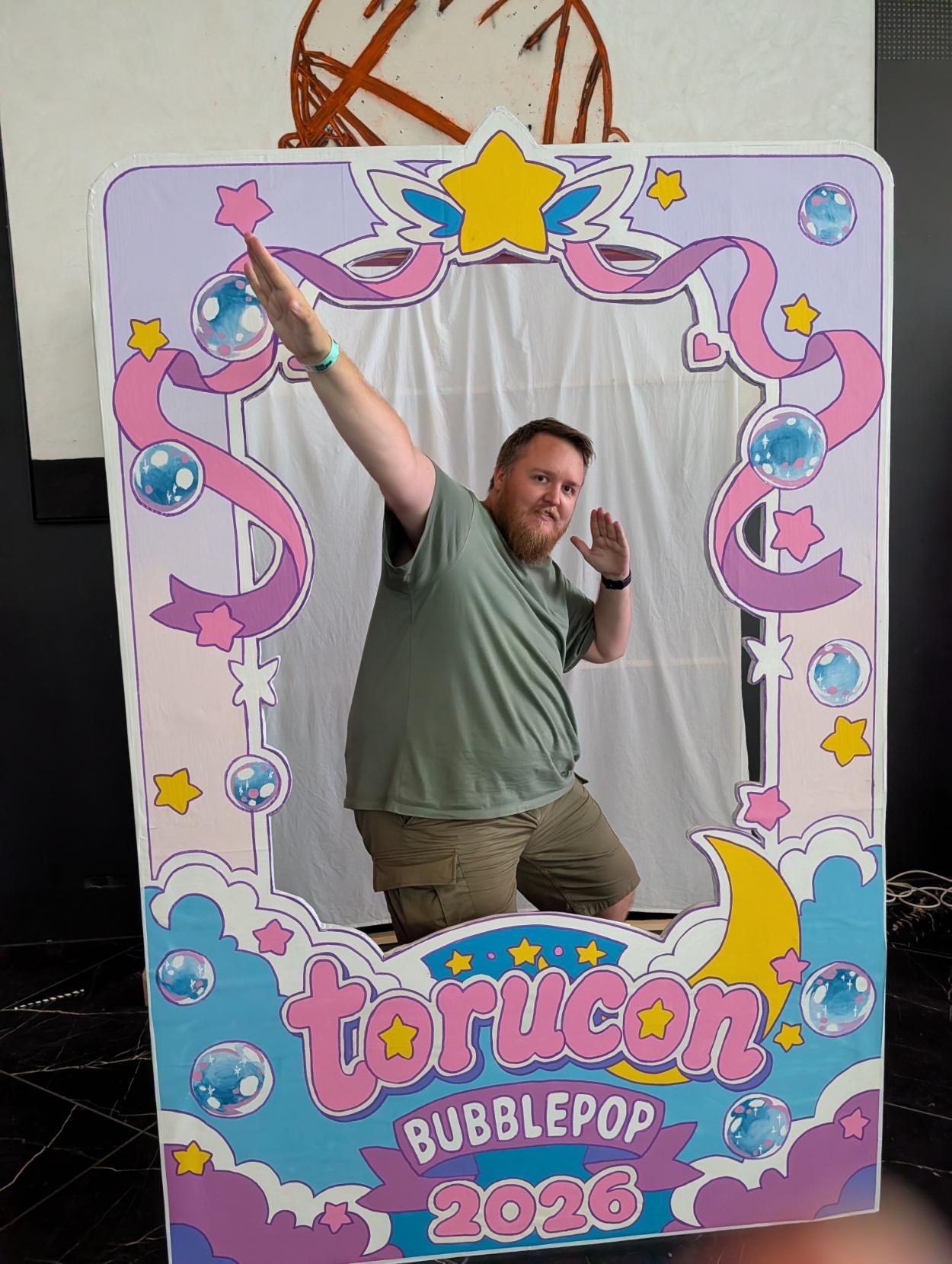 Ein mann med skjegg, grøn t-skjorte og shorts poserer dynamisk inni ein fargerik, dekorert fotoramme på Torucon. Han strekkjer den eine armen skrått oppover og den andre ut til sida. Den pastellfarga ramma er pynta med rosa band, stjerner, såpebobler og ein gul månesigd. Nederst på ramma står teksten «Torucon», «Bubblepop» og «2026» i spretten, fargerik skrift på ein bakgrunn av lyseblå og lilla skyer.