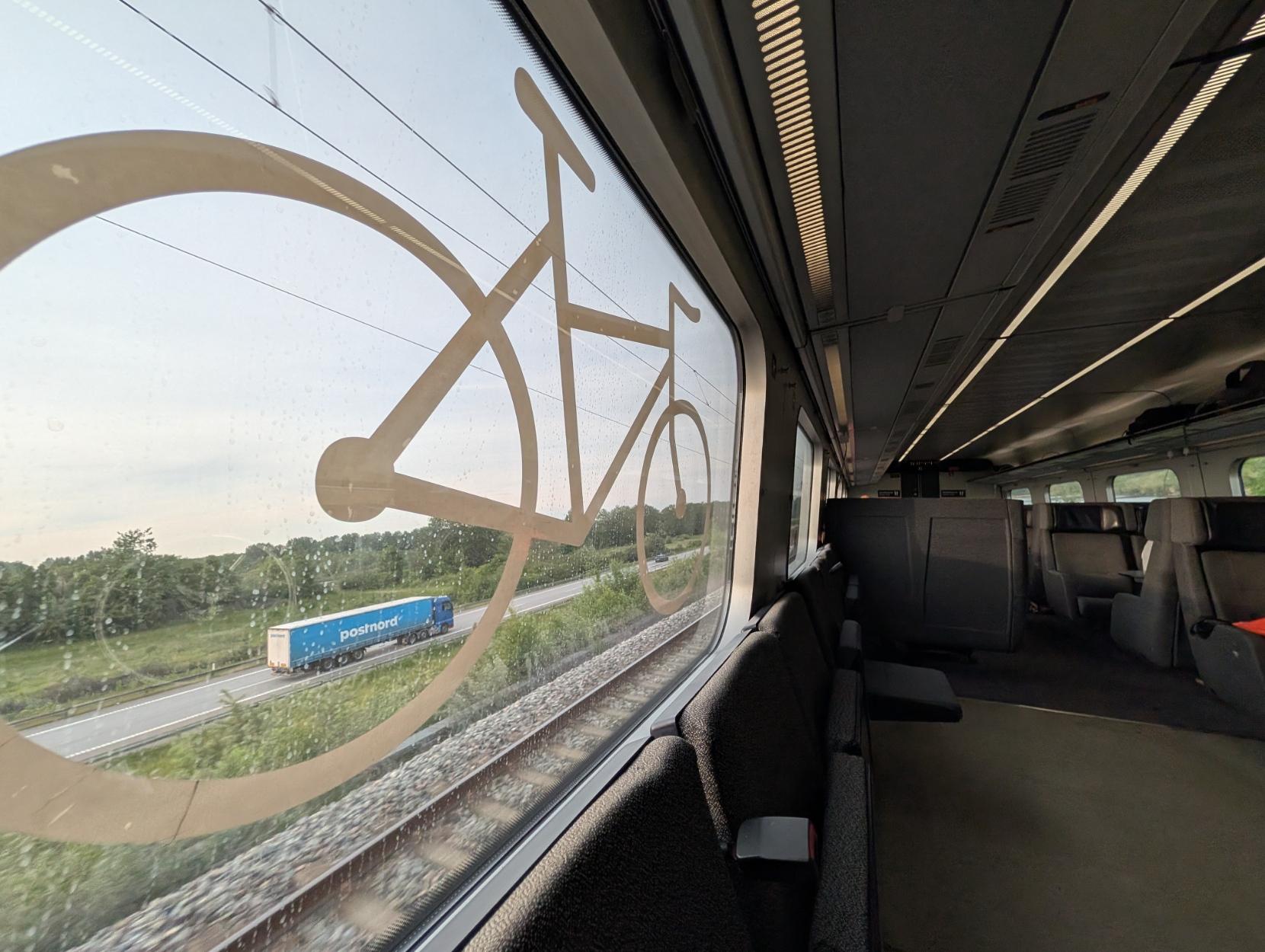 Eit bilete innanfrå fleirbrukskupeen på eit elektrisk DSB IR4-tog i fart. Til venstre dominerer eit stort panorama-vindauge med eit gigantisk, lyst sykkelsymbol klistra over glasset. Gjennom det småregna vindauget ser ein ut på ein motorveg som går parallelt med jernbanesporet, der ein lyseblå PostNord-lastebil køyrer av garde. Landskapet utanfor er prega av grøne sletter og tre under ein ljos kveldshimmel. Inne i toget ser ein ei rekkje med mørkegrå klappsete langs vindauget, eit opent golvareal for syklar eller barnevogner, og vanlege passasjersete lenger bak i den dagslyste kupéen.