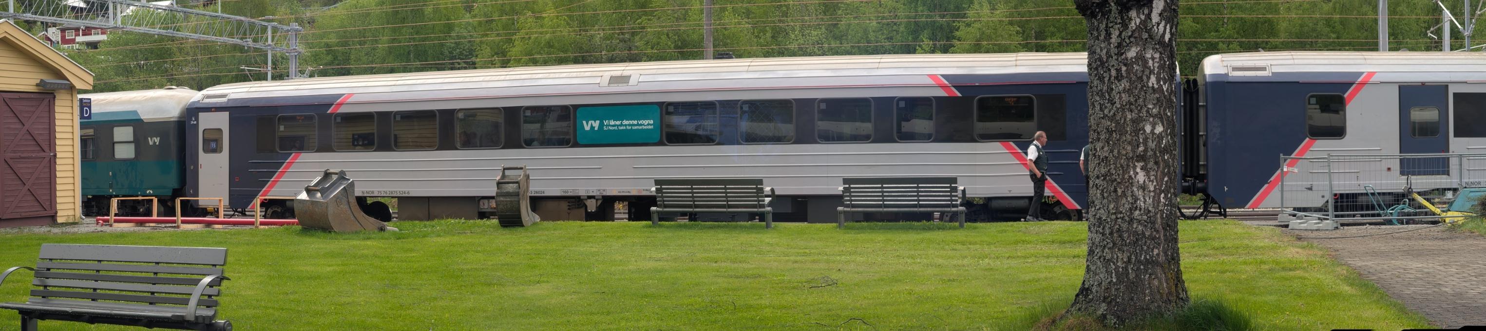 Eit nærbilete i breiddeformat som viser ei sølvgrå og mørkeblå Type 7-passasjervogn (merka B7-6 26024) på eit Vy-tog på Ål stasjon. Vognen har ein stor, grøn tekstellips som seier «Vy – Vi låner denne vogna. SJ Nord, takk for samarbeidet». Til venstre er ein mørkegrøn vogn ende i syne. Framfor toget er det ein velstelt grøn plen med to parkbenkar og to gravemaskinskuffer som ligg på bakken. Til høgre står ein togkonduktør i uniform ved sidan av stammen til eit stort tre, og i bakgrunnen anar ein grøne skogkledde åsar.