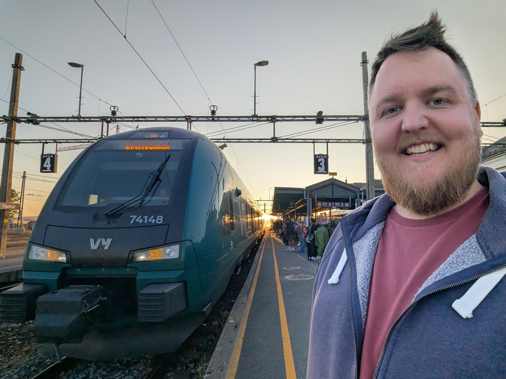 Ein selfie av ein breitt smilande mann med skjegg, vinraud t-skjorte og blå hettejakke på ein perrong på Hamar stasjon i solnedgangen. Til venstre står fronten på eit grønt og mørkegrått elektrisk Vy-tog av typen Stadler FLIRT (Vy litra BM74), merka med settnummeret «74148» under frontruta og Vy sin kvite logo på snuten. Destinasjonsskjermen over frontruta viser «RE10 Drammen», og toget går sørover om Oslo S. Frontlysa og dei oransje blinklysa lyser. I bakgrunnen heng skilta «SPOR 4» og «SPOR 3» under køyreleidningane, folk ventar bortover perrongen ved stasjonsbygget, og den låge sola står som ein oransje glød mellom toga under ein ljos, klår kveldshimmel.

🚆 Tog: Type 74 (FLIRT) · Litra: BM74 · Nr: 74148 · Stasjon: Hamar · Bane: RE10 · Selskap: Vy