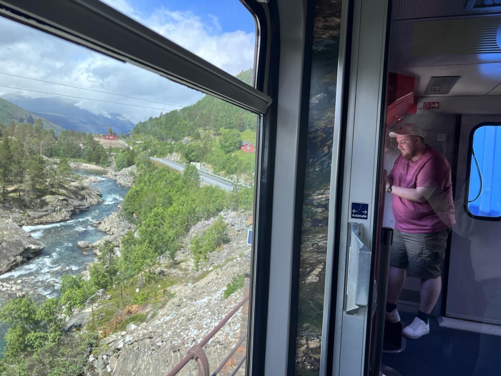 Utsyn gjennom eit ope vindauge om bord på The Golden Train (Raumabana), der det dramatiske norske landskapet opnar seg. Til venstre ser ein den klåre, skumgitte elva Rauma som renn gjennom eit trongt gjel omkransa av grøne furutre, bergveggar og bratte, skogkledde åssider under ein delvis blå og skyete himmel. Til høgre i biletet ser ein ein refleksjon i vindaugsglaset til ei innvendig dør, der ein mann med skjegg, ljos caps, rosa t-skjorte og shorts står inne i den moderne togvogna.