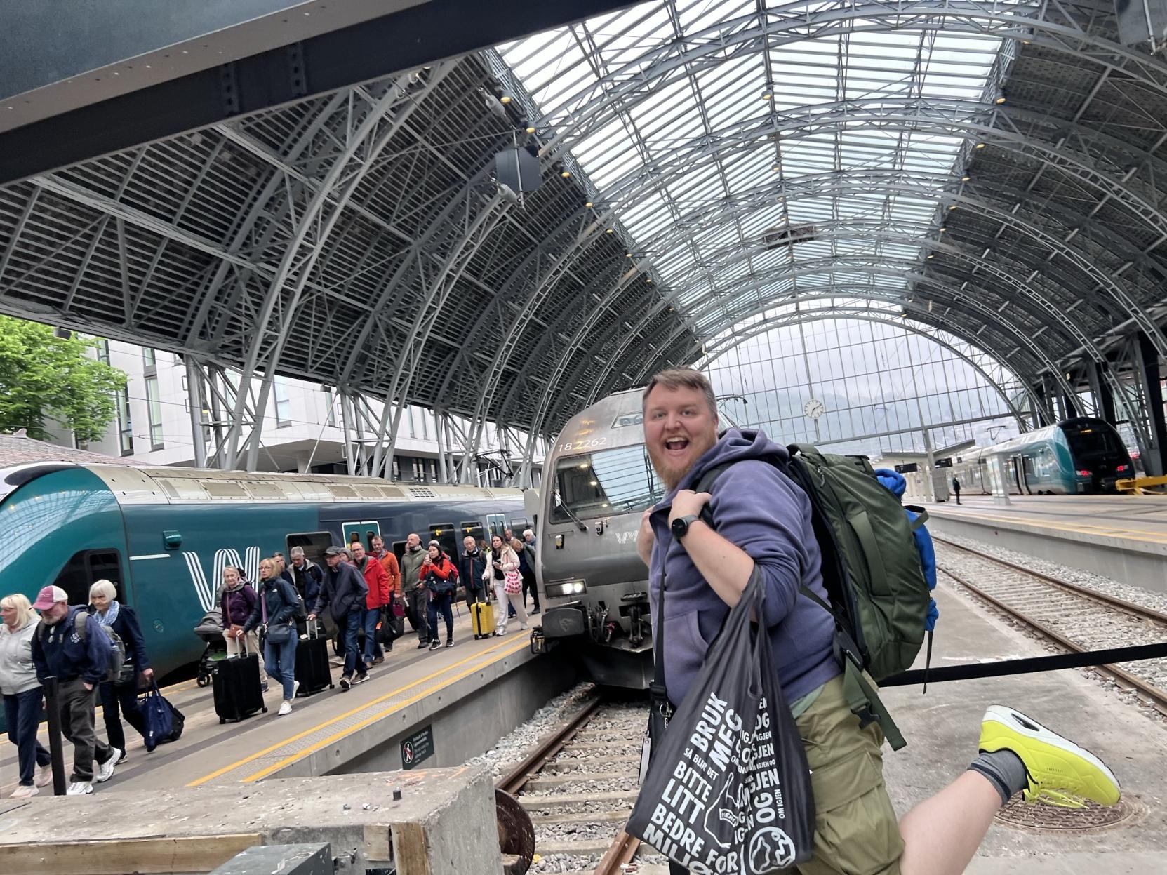 Ein energisk og morosam selfie av ein mann med skjegg, lilla hettegenser og ein stor, grøn ryggsekk, som gjer eit hopp på ein togperrong inne på Bergen stasjon. Han held ein gjenbrukbar handlepose i den eine handa og smiler breitt til kameraet med ein fot løfta bak seg. Sentralt i bakgrunnen står lokomotivet han kom med, det mørkegrå elektriske Vy-lokomotivet av typen El 18 (nummer 18 2262). Til venstre strøymer passasjerar med trillekoffertar ut frå dei ljosare grøne og mørkeblå Vy-vognene til Bergensbanen. Heilt til høgre på naboperrongen står eit anna grønt Vy-tog parkert. Over heile stasjonsområdet breier den veldige og arkitektoniske bogehalden i stål og glas seg ut.