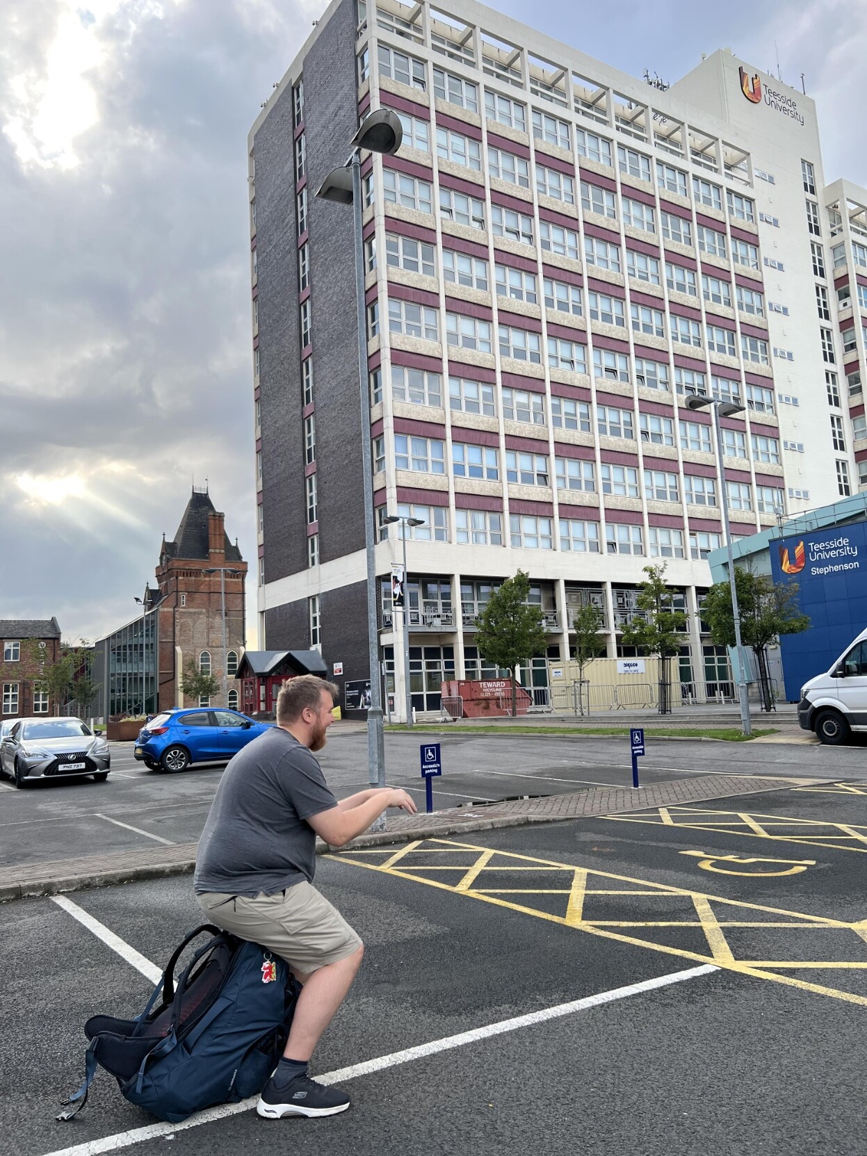 @markus@skvip.lol sitt på ein ryggsekk og latar som om han er på ein datamaskin. Han sitt midt på ein parkeringsplass med ei høgblokn i bakgrunnen. Høblokka har logoen til «Teesside University».