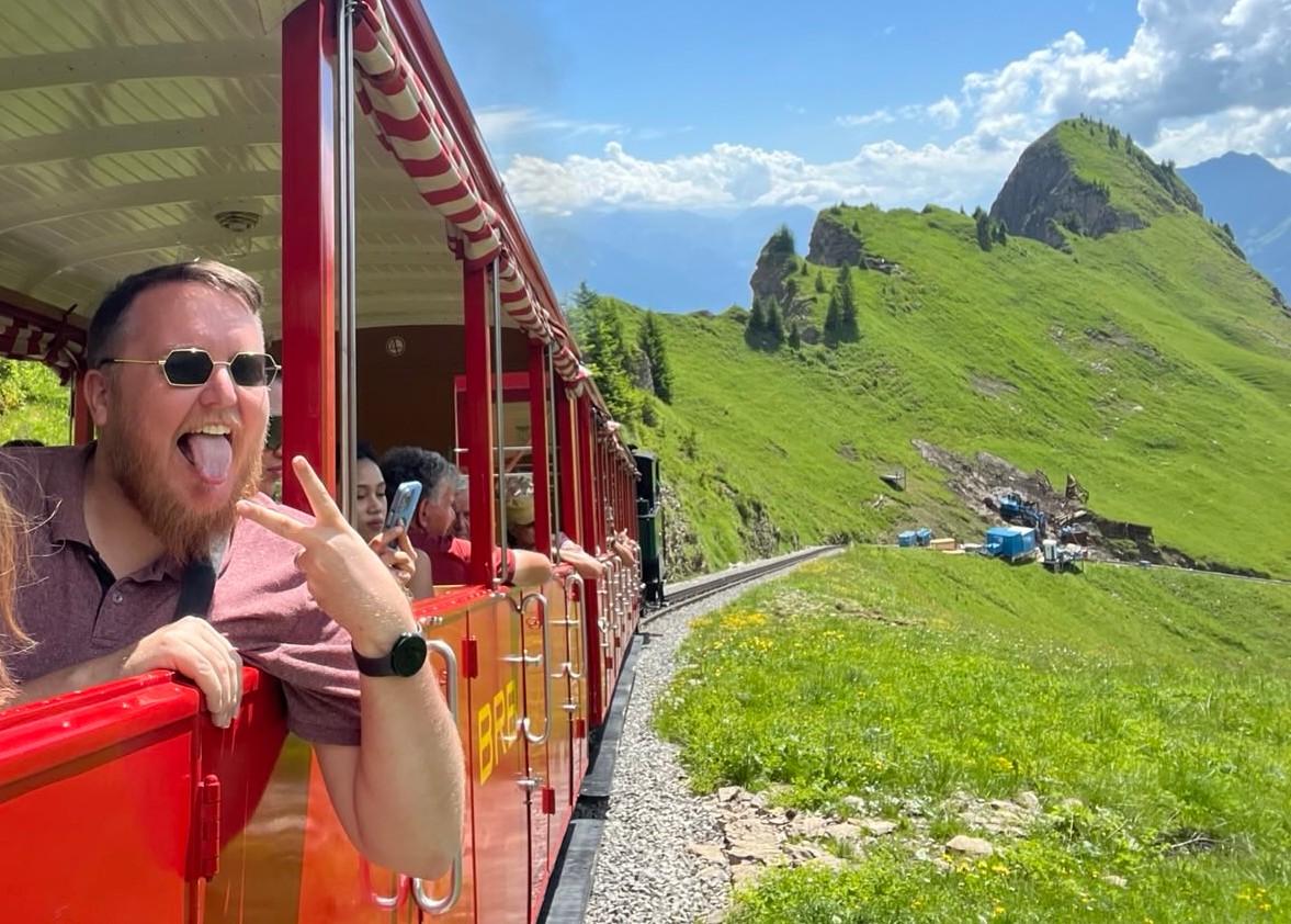 Bilete av mann (@markus@skvip.lol) i open togvogn. I bakgrunnen er det eit veldig grønt fjell og blå himmel